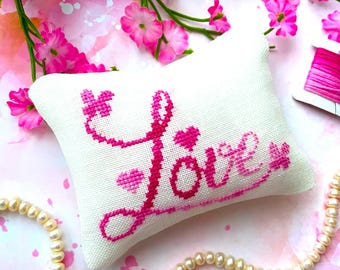 Puede incluir: Una almohada blanca con bordado rosa que dice "Love" con un collar de perlas rosas y tijeras doradas sobre un fondo floral rosa.