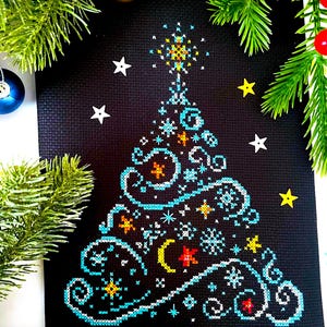 Könnte beinhalten: Ein schwarzes Stoffstück mit einem Kreuzstichmuster eines Weihnachtsbaums. Der Baum besteht aus hellblauen Schnörkeln, weißen Schneeflocken sowie kleinen orangefarbenen und gelben Sternen. Ein gelber Halbmond und ein Stern sind enthalten.