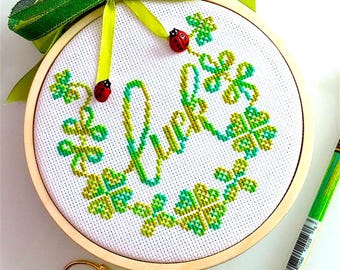 Könnte beinhalten: Ein weißer Stickrahmen mit einem grünen und gelben Kreuzstich-Design aus vierblättrigen Kleeblättern und dem Wort "Luck" in einer kursivschriftlichen Schriftart. Zwei rote Marienkäfer sind auf das Design gestickt. Der Rahmen ist mit einem grünen Band gebunden.
