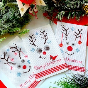 May include: Three handmade Christmas cards with cross-stitched reindeer faces, red noses, and the words "Merry Christmas." Each card has a red and white checked ribbon. Festive greenery and ornaments surround the cards.