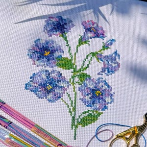 Könnte beinhalten: Kreuzstich-Stickerei mit blauen und violetten Blumen auf weißem Stoff. Grüne Stiele und Blätter sind sichtbar. Bunte Stickgarne und eine goldene Schere im Vordergrund.