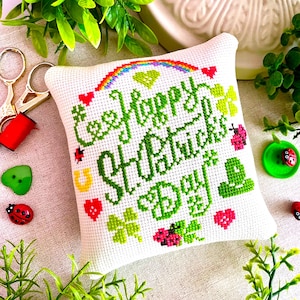Puede incluir: Un cojín blanco bordado con "Happy St. Patrick's Day" en verde. El diseño incluye un arcoíris, tréboles, corazones y mariquitas. Rodeado de follaje verde y elementos decorativos.