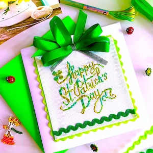 May include: A handmade St. Patrick's Day card with a green scalloped border and a large green bow. The card features cross-stitch embroidery with the words "Happy St. Patrick's Day" in green and gold thread. Additional craft supplies include scissors, ribbon, and thread.
