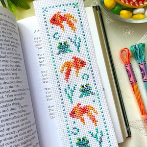 Könnte beinhalten: Handgefertigter Kreuzstich-Lesezeichen mit einem Design aus orangefarbenen Fischen und grünen Fröschen zwischen Wasserpflanzen. Das Lesezeichen befindet sich in einem offenen Buch. Buntes Stickgarn und dekorative Steine sind ebenfalls sichtbar.