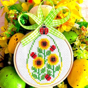 Puede incluir: Un aro de tela blanco con un diseño de punto de cruz de una planta verde con tres girasoles amarillos y mariquitas rojas. El aro está rodeado de decoraciones de Pascua amarillas y verdes, incluyendo un pollito blanco, flores amarillas y huevos moteados.