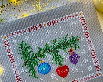 May include: Cross-stitch embroidery of a Christmas tree with ornaments that say "Peace", "Love", and "Joy". The background is a light gray with a white snowflake pattern. The border is red and white with the words "Peace, Love & Joy" repeated around the edge.