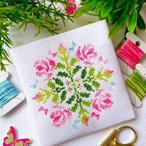 May include: A white cross-stitch embroidery featuring a floral design with pink and green flowers. The embroidery is framed by a white border and is surrounded by embroidery floss and scissors.