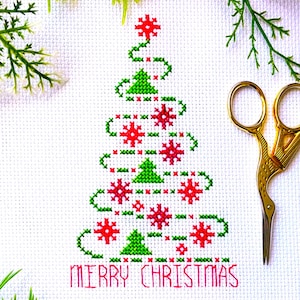 May include: A white fabric piece featuring a cross-stitch design of a Christmas tree composed of green triangles and red snowflake patterns, with the text MERRY CHRISTMAS stitched in red below the tree and gold-colored metal scissors nearby.