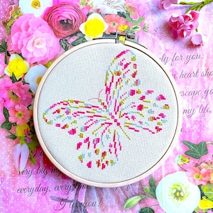 May include: A cross-stitch butterfly design in a wooden embroidery hoop. The butterfly is stitched with pink, yellow, and blue thread on a light-coloured fabric. The background is a pink floral design with text that reads "very big my everyday, every time I love you!"
