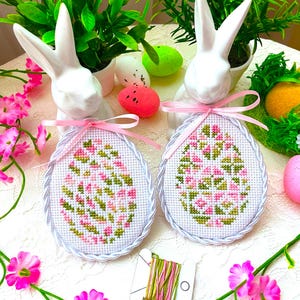 Könnte beinhalten: Zwei weiße Keramik-Hasenfiguren, die rosa und weiße bestickte Ostereier mit grünen und rosa Blumenmustern halten. Die Eier sind von rosa Blumen und grünem Laub umgeben.