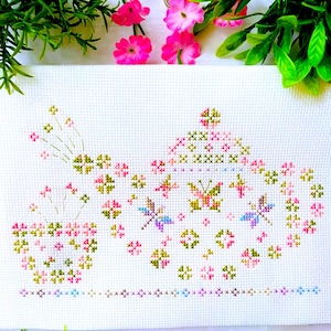 May include: A white fabric piece featuring a cross-stitch pattern of a teapot and a teacup. The design is composed of small, colorful X-shaped stitches in shades of pink, green, and blue, depicting floral motifs and delicate dragonfly figures.