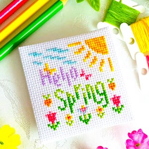 Puede incluir: Un cuadrado de punto de cruz blanco con las palabras "Hello Spring" en morado y verde, con un sol, flores y un cielo azul. Lápices de colores y carretes de hilo de varios colores están dispuestos alrededor del cuadrado.