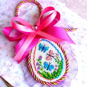 May include: A pink and gold Easter egg ornament featuring a cross stitch design of butterflies and flowers. The egg is adorned with a pink ribbon and a gold cord.
