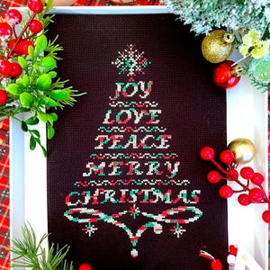 May include: A framed cross-stitch artwork with a Christmas tree design. The tree is made up of the words "JOY," "LOVE," "PEACE," "MERRY," and "CHRISTMAS" in white, red, and green thread on a dark brown background. The artwork is surrounded by festive decorations.