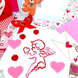 May include: A white cross-stitch with a red embroidered image of a cupid holding a heart. The cross-stitch is surrounded by pink and red felt hearts.