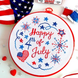 May include: A white embroidery hoop with red and blue embroidery depicting fireworks, stars, and the text "Happy 4th of July".