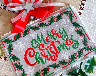 May include: A white cross-stitch with a red and green holly border and the words "Merry Christmas" in red and green lettering. The cross-stitch is on a white background with a white border and a white pom-pom trim.