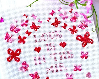 Puede incluir: Bordado blanco de punto de cruz con una forma de corazón rojo hecha de mariposas rosas y rojas. El texto "LOVE IS IN THE AIR" está bordado en rojo dentro del corazón.