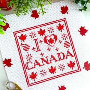 Peut inclure: Tissu blanc brodé au point de croix avec un motif rouge disant « I CANADA » avec un cœur et une feuille d'érable. Des découpes de feuilles d'érable rouges et des ciseaux dorés sont éparpillés autour du tissu. Fond vert et rouge.