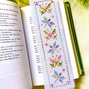 May include: A handmade white cross-stitch bookmark featuring a repeating butterfly design in shades of pink, green, blue, and yellow. The bookmark is placed in an open book with text visible on the pages. The book has a green spine.