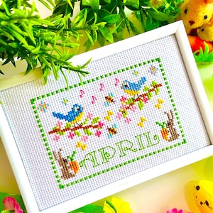 Peut inclure: Une broderie au point de croix encadrée avec un cadre blanc. Le motif représente deux oiseaux bleus perchés sur une branche fleurie, avec le mot "APRIL" brodé en vert. Inclut des abeilles et des lapins.