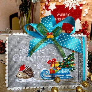 Puede incluir: Un diseño de punto de cruz gris y blanco con un erizo tirando de un trineo con un árbol de Navidad y regalos. El diseño está enmarcado con un borde blanco y una cinta azul con lunares blancos.