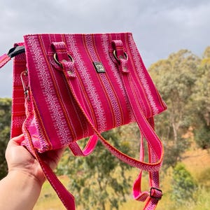 May include: A pink handbag with a woven pattern and a shoulder strap. The bag has a rectangular shape with a zip closure and metal hardware. The strap is adjustable and the bag is held up by a hand.