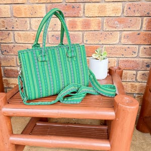 May include: A green striped tote bag with a top handle and a long shoulder strap. The bag is sitting on a wooden table.