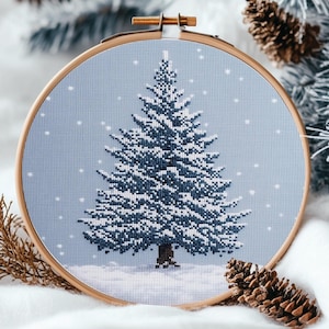 Könnte beinhalten: Eine blaue und weiße Kreuzstich-Stickerei eines schneebedeckten Kiefernbaums in einem hölzernen Stickrahmen.