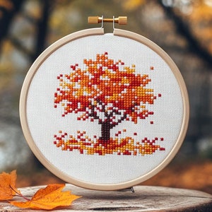May include: A wooden embroidery hoop with a cross-stitch design of an orange, yellow, and red tree. The tree is surrounded by smaller orange, yellow, and red leaves.