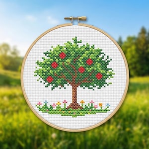 Puede incluir: Bordado de punto de cruz de un manzano con manzanas rojas, hojas verdes y un tronco marr&oacute;n. El &aacute;rbol est&aacute; sobre un fondo blanco dentro de un bastidor de bordado de madera. Peque&ntilde;as flores y hierba en la base del &aacute;rbol.