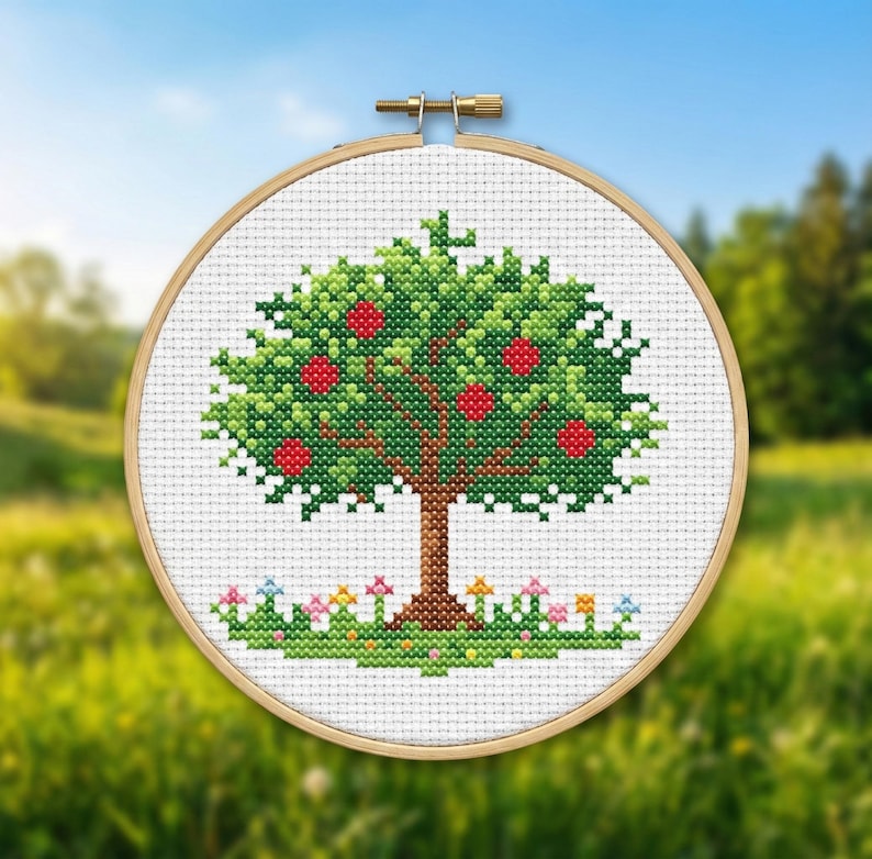 Puede incluir: Bordado de punto de cruz de un manzano con manzanas rojas, hojas verdes y un tronco marr&oacute;n. El &aacute;rbol est&aacute; sobre un fondo blanco dentro de un bastidor de bordado de madera. Peque&ntilde;as flores y hierba en la base del &aacute;rbol.