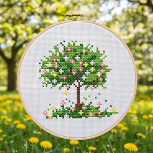 Puede incluir: Un bordado de punto de cruz de un &aacute;rbol en un aro de madera. El &aacute;rbol tiene un dosel verde con flores rosas, amarillas y blancas. El tronco es marr&oacute;n y la hierba en la base tiene flores similares. El fondo es verde borroso.