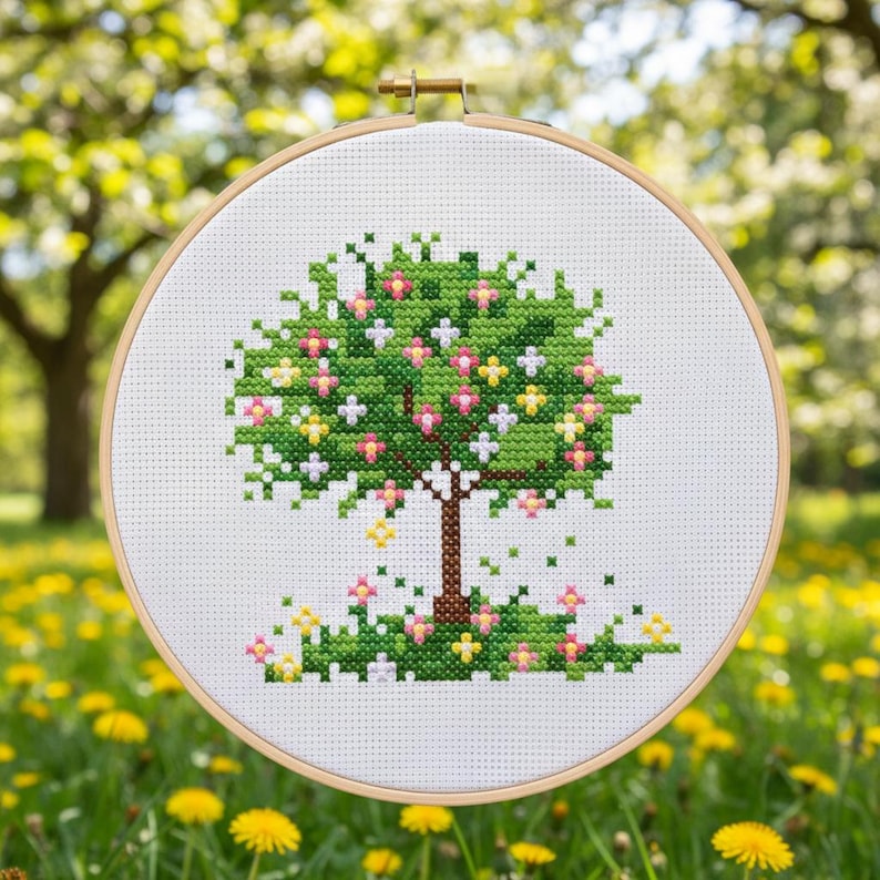 Puede incluir: Un bordado de punto de cruz de un &aacute;rbol en un aro de madera. El &aacute;rbol tiene un dosel verde con flores rosas, amarillas y blancas. El tronco es marr&oacute;n y la hierba en la base tiene flores similares. El fondo es verde borroso.