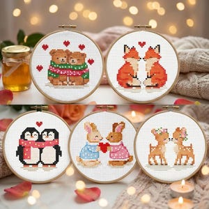 Peut inclure: Cinq cercles de broderie au point de croix représentant des couples d'animaux : ours, renards, pingouins, lapins et cerfs. Chaque motif comprend des cœurs et est posé sur un fond blanc. Les cercles sont encadrés dans des cerceaux à broder en bois.