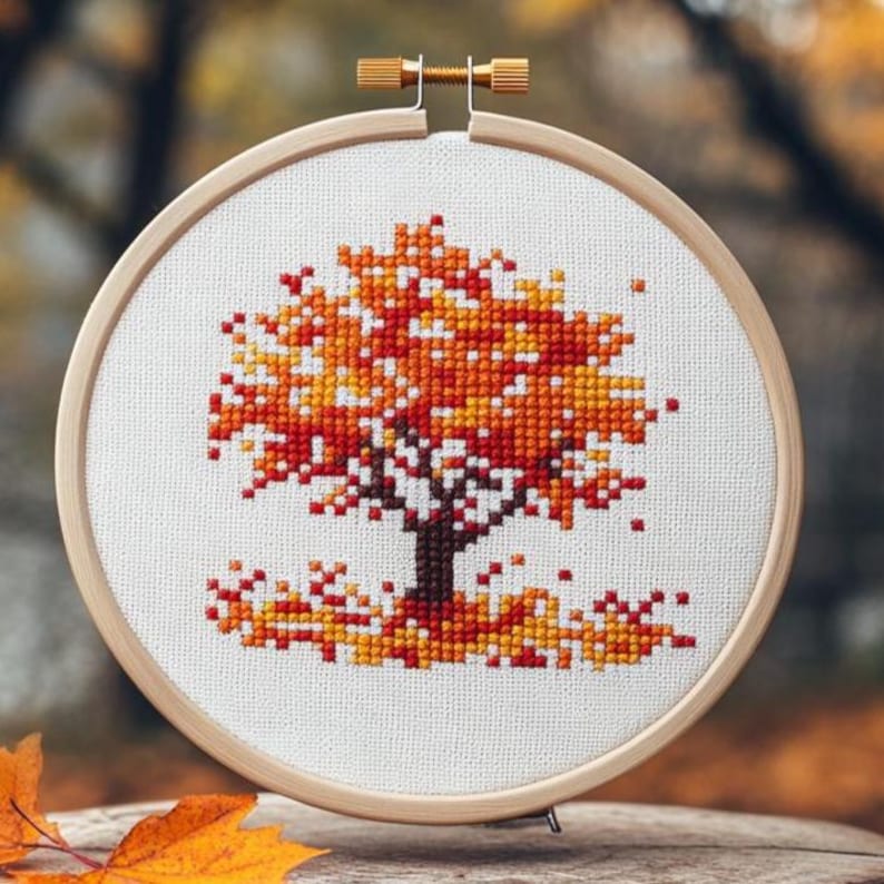 Puede incluir: Bordado de punto de cruz de un &aacute;rbol oto&ntilde;al, en un bastidor de madera. El &aacute;rbol tiene hojas naranjas y amarillas, un tronco marr&oacute;n y hojas ca&iacute;das en la base. El fondo est&aacute; borroso.
