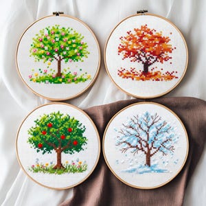 Op de afbeelding: Vier borduurringen met kruissteek die bomen in verschillende seizoenen uitbeelden: lente, herfst, zomer en winter. Elke boom is weergegeven in levendige kleuren, met details zoals bloemen, bladeren en sneeuw. De ringen staan tegen een witte en bruine achtergrond.