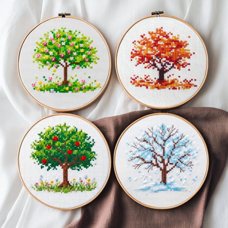 Puede incluir: Cuatro aros de bordado de punto de cruz que representan &aacute;rboles en diferentes estaciones: primavera, oto&ntilde;o, verano e invierno. Cada &aacute;rbol est&aacute; representado en colores vibrantes, con detalles como flores, hojas y nieve. Los aros est&aacute;n sobre un fondo blanco y marr&oacute;n.