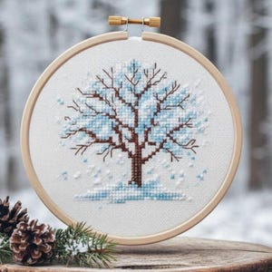Puede incluir: Bordado de punto de cruz de un &aacute;rbol de invierno en un aro de madera. El &aacute;rbol presenta un tronco y ramas marrones con hojas azul claro y nieve cayendo. El bordado est&aacute; sobre un fondo borroso de bosque invernal.