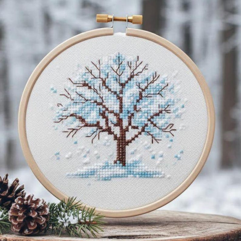 Puede incluir: Bordado de punto de cruz de un &aacute;rbol de invierno en un aro de madera. El &aacute;rbol presenta un tronco y ramas marrones con hojas azul claro y nieve cayendo. El bordado est&aacute; sobre un fondo borroso de bosque invernal.