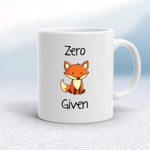 May include: White ceramic mug featuring a cartoon fox. The fox is orange and brown, with the words "Zero Given" in black text above and below the fox. The mug has a curved handle.