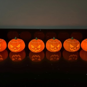 Puede incluir: Seis calabazas naranjas en forma de jack-o'-lantern con caras talladas están iluminadas desde el interior, creando un brillo cálido. Las calabazas están dispuestas en una fila sobre una superficie negra.