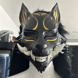 May include: A black wolf mask with gold accents and white fangs. The mask has a mesh eye covering and is surrounded by black faux fur. The design features stylized curves and lines, creating a fierce and detailed appearance.