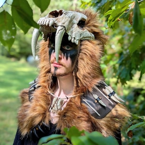 May include: A costume headpiece featuring a realistic sabre-toothed tiger skull with long white fangs, integrated into a thick brown faux fur hood. The accessory includes a dark leather shoulder mantle adorned with multiple white bone-like spikes.