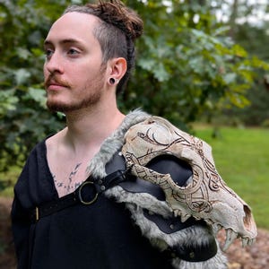 May include: A shoulder armour piece featuring a carved, off-white animal skull with intricate knotwork patterns. The skull is mounted on a black leather base with grey faux fur trim, secured by black leather straps and a metal ring across the chest.