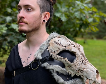 May include: A shoulder armor piece featuring a sculpted, off-white animal skull with intricate dark brown etched knotwork patterns. The skull is mounted on a black leather base with grey faux fur trim, secured by black leather straps and a metal ring.