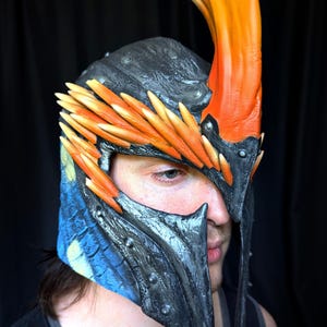May include: A fantasy helmet with a black, blue, and orange color scheme. The helmet features a large, curved orange horn and orange feather-like accents. The face guard is dark gray with riveted details, and the helmet has a textured appearance.