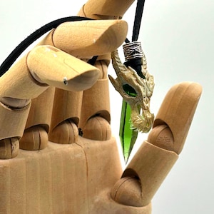 Puede incluir: Un colgante de cristal verde con una cabeza de dragón esculpida en la parte superior. El colgante está unido a un cordón negro con una cuenta de plata.