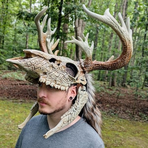 May include: A wearable deer skull mask featuring intricate Celtic-style knotwork carvings across the bone surface. The mask includes large, textured antlers with brown and cream coloration, and a long, faux fur mane attached to the back.