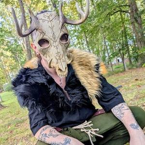 May include: A beige skull mask with large antlers and carved markings. The mask is worn with a black top and a faux fur shoulder piece. The person is wearing green trousers and has tattoos on their arms.